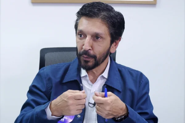 Imagem colorida mostra Ricardo Nunes, homem branco, de cabelo e barba pretos, vestindo jaqueta azul e camisa branca, sentado em uma cadeira com uma parede branca ao fundo, conversando - Metrópoles