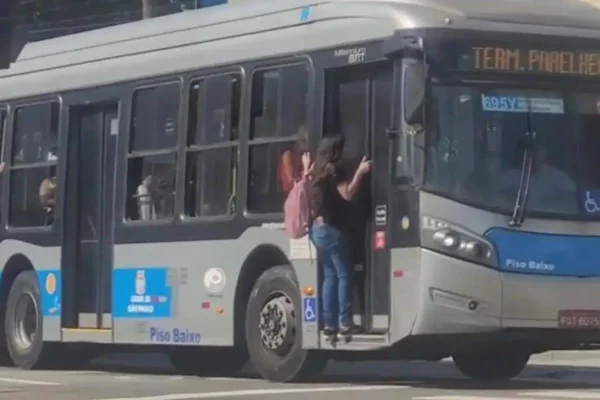 Imagem colorida mostra mulher pendurada na porta de um ônibus em São Paulo - Metrópoles