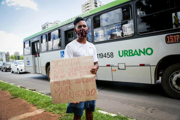 Imagem colorida de um homem negro, vestindo camiseta branca e bermuda jeans, segurando um cartaz escrito: "Ajuda, sou venezuelano, tô na rua. Pode me dar qualquer valor. Obrigado."