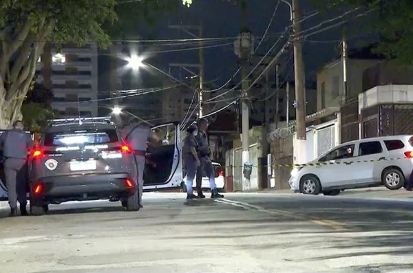 Imagem colorida. Rua a noite com carro da polícia estacionado enquanto dois policiais e uma policial analisam área isolada.