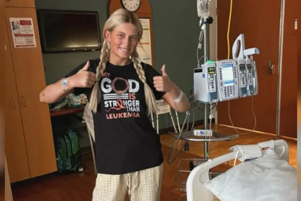 Foto colorida de jovem com cabelos longos e loiros, blusa preta, fazendo sinal positivo em ambiente hospitalar-Metrópoles.
