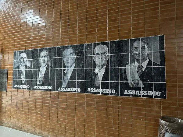 Fotografia colorida de mural com fotos de homens em preto e branco e palavra "assassino"