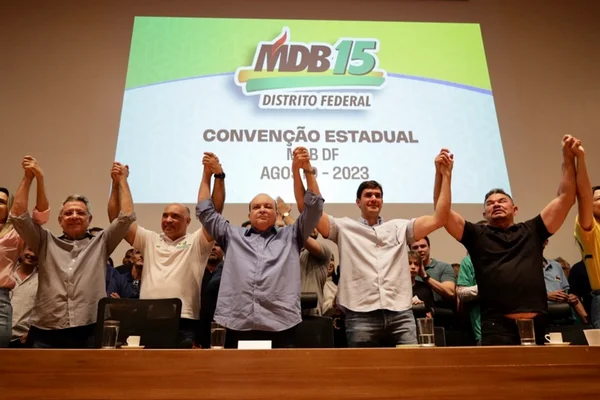 Foto colorida de sete pessoas, com as mãos unidas e os braços para cima; entre elas, há o governador Ibaneis Rocha, ao centro, além dos deputados distritais Wellington Luiz e Hermeto; ao fundo, há uma projeção com informações sobre a convenção estadual do MDB-DF