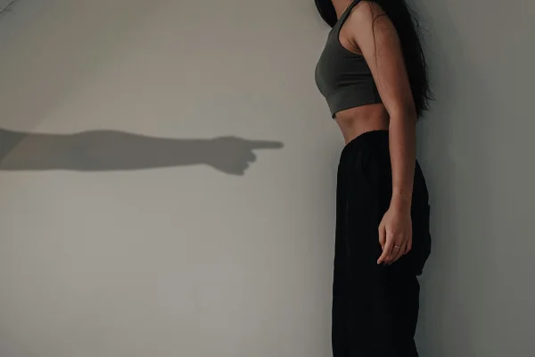 Foto colorida de mulher esguiando a barriga em frente a uma sombra de um braço apontado o dedo para ela - Metrópoles.