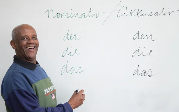 Foto colorida de homem negro, sorrindo e segurando uma caneta, em frente a uma lousa, com palavras em alemão - Metrópoles