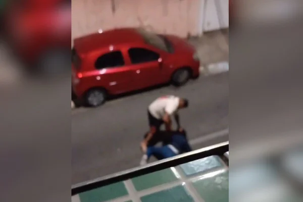 imagem colorida mostra homem de camiseta branca se projetando sobre mulher caída na rua. ele segura a cabeça dela no chão de forma violenta