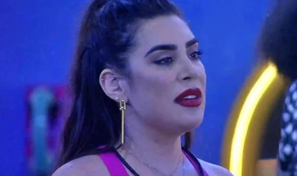 Na imagem colorida, uma mulher está posicionada no centro. Ela tem cabelos curtos e loiros, olhos castanhos e olha seriamente para a câmera