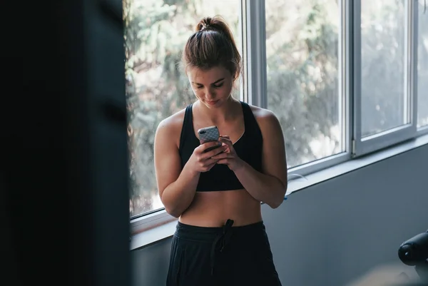 Foto mostra mulher fitness, na academia, mexendo no celular. Vendo postagens fitness. Metrópoles