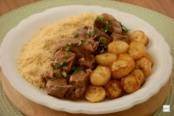 Picadinho de carne refrogado com banana da terra e farofa em prato branco e sousplat verde - Metrópoles