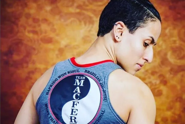 Em foto colorida atleta de cabelo curto está de costas usando camiseta regata azul na qual está escrico Macfer em meio a um circulo do ying yang, branco e azul, com um fundo semelhante a chamas de fogo - Metrópoles