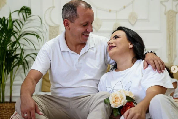 Foto colorida de homem (esq) sorrindo para mulher que o olha, com semblante apaixonado. Ambos vestem roupas brancas - Metrópoles
