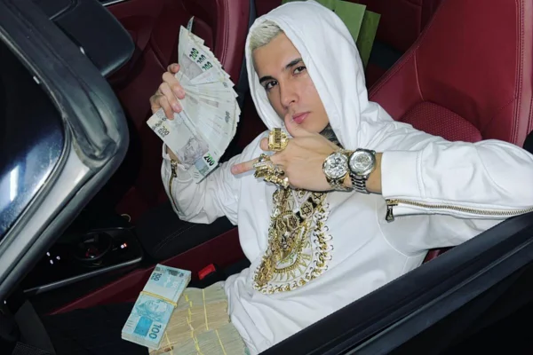 Foto colorida de jovem branco, com moletom branco, cujo capuz cobre-lhe os cabelos descoloridos, segurando notas de dinheiro e ostentando anéis dourados, enquanto ocupa bando do motorista de carro de luxo conversível - Metrópoles
