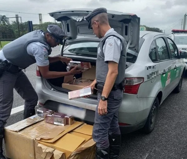 Dois agentes com fardas cinzas recolhem caixas de cigarros de porta-malas de carro sedan prata - Metrópoles