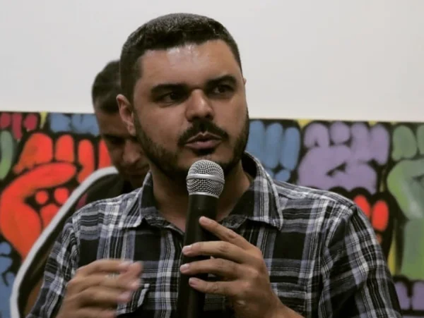 Imagem colorida mostra Laercio Ribeiro, homem branco, de cabelos pretos, camisa xadrez, falando ao microfone na frente de um fundo com uma parede com grafites - Metrópoles
