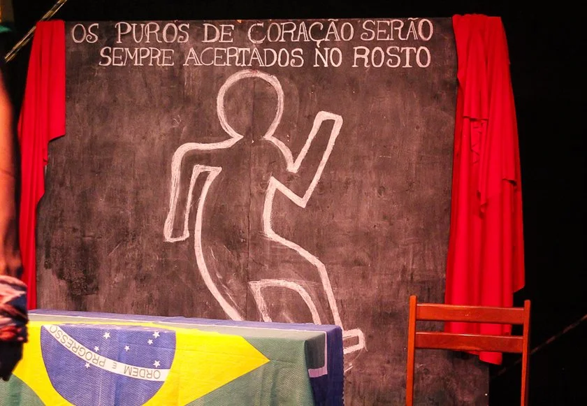 O cenário com a frase "Os puros de coração serão sempre acertados no rosto" permanece no palco do início ao fim O cenário com a frase "Os puros de coração serão sempre acertados no rosto" permanece no palco do início ao fim