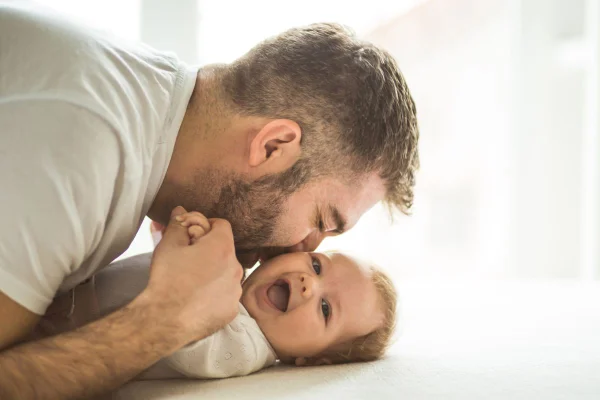 Pai beijando rosto de bebê, que olha para a câmera sorrindo