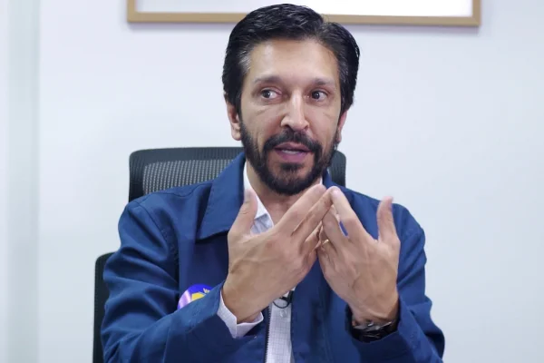Imagem colorida mostra Ricardo Nunes, homem branco, de cabelo e barba pretos, vestindo jaqueta azul e camisa branca, sentado em uma cadeira com uma parede branca ao fundo, conversando - Metrópoles