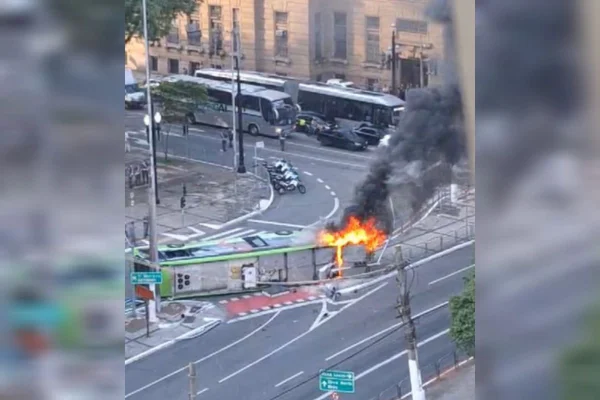 ônibus verde está tombado atravessado em rua, com chamas e fumaça saindo da parte dianteira - Metrópoles