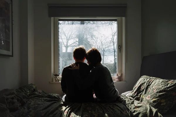 Imagem colorida mostra duas pessoas sentadas e abraçadas em cima de uma cama dentro de um quarto iluminado pelo sol - Metrópoles
