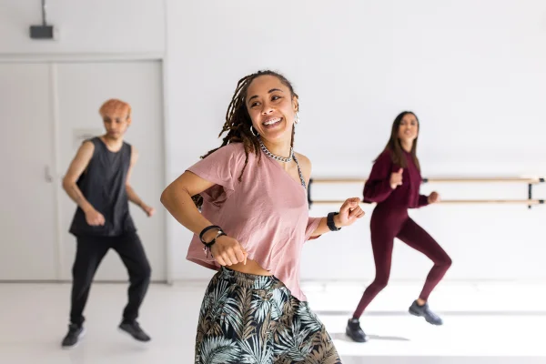 Três pessoas sorridentes dançando em sala branca - Metrópoles