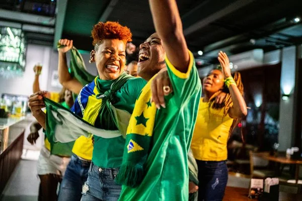 Torcedores comemoram gol do Brasil em um bar. Eles estão vestidos com as cores da bandeira brasileira. Dicas de receita para a Copa do Mundo 2022