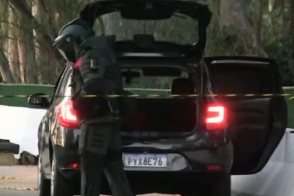 Imagem colorida de agente com roupa anti bombas vendo carro preto com porta-malas aberto. Metrópoles