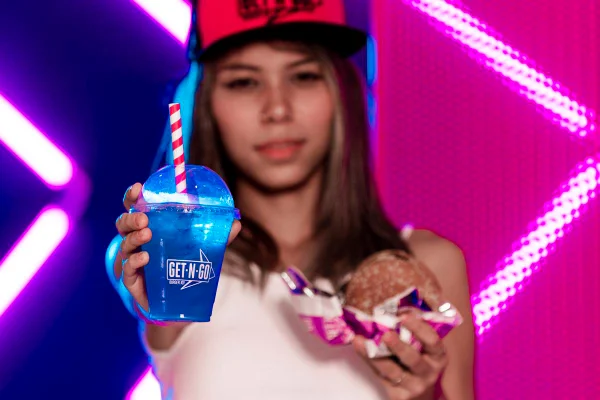 foto de menina desfocada com copo transparente em primeiro plano com liquido azul e segurando hamburguer na outra mão