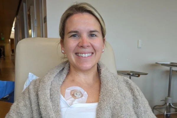 Foto colorida de mulher em ambiente hospitalar, sentada e sorrindo para a câmera. Ela tem um curativo no peito - Metrópoles