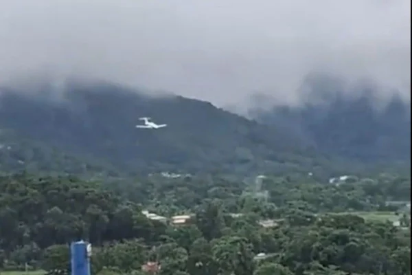avião plana entre nuvens cinzentas e chuvas; árvores e casas compõem o plano de fundo - Metrópoles