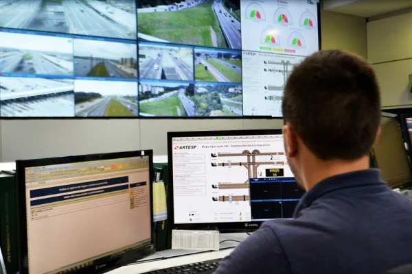 Imagem colorida mostra homem sentado, de costas. Ele está virado para uma central com dois monitores de computadores e uma tela grande na parede - Metrópoles