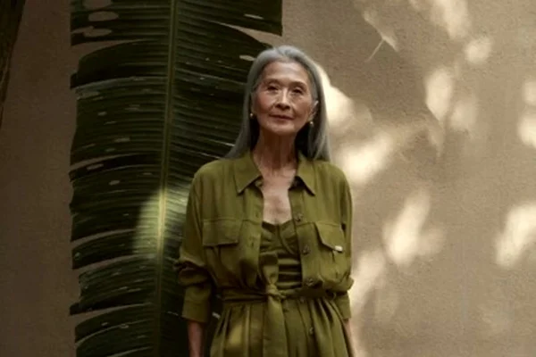 Modelo idosa posa com look verde e folha grande de planta no fundo - Metrópoles