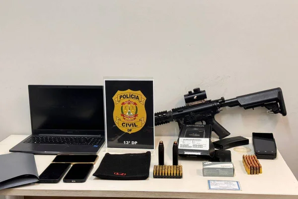 Foto colorida de computador, celulares, fuzil e muniçoes em cima de mesa