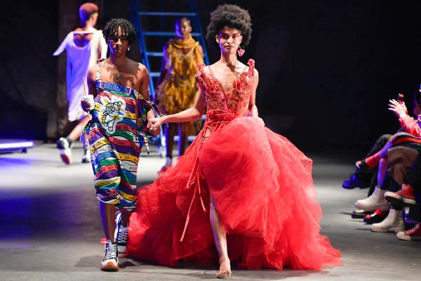 Em passarela de moda, modelos de mãos dadas. Um dos looks é um vestido vermelho volumoso e o outro é uma espécie de macacão colorido - Metrópoles