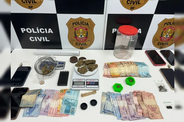 Imagem colorida de uma mesa com notas em espécie, porções de drogas, uma balança de precisão e vários celulares; ao fundo, um backdrop da Polícia Civil do DF