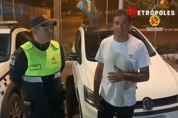 Policial militar com farda verde fluorescente e azul-escuro, usando boné azul-escuro da corporação, ao lado de homem pardo de camiseta branca, come em frente a carro da PMDF e a um Volkswagen branco
