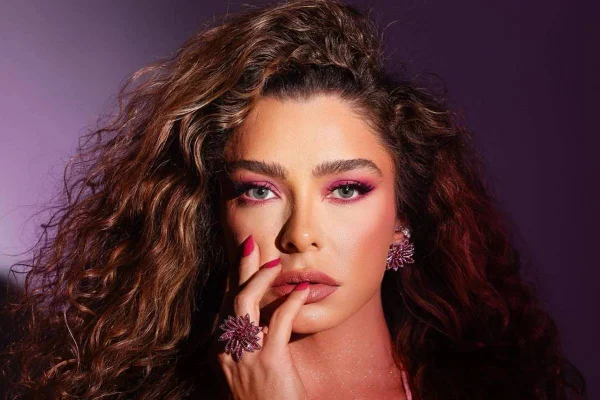Foto colorida de Fabíola de Andrade com a mão no rosto, em fundo roxo - Metrópoles