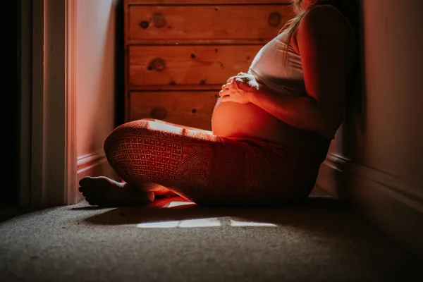 Foto colorida em tons de vermelho, de mulher grávida sentada no chão sob luz solar. - Metrópoles.