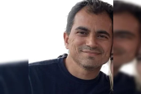 Foto colorida de homem branco, de cabelos escuros e vestido com camiseta preta, com fundo branco