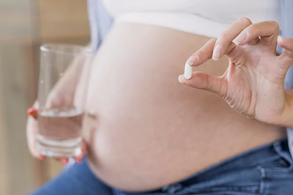 Foto colorida de mulher grávida segurando um copod'água e uma cápsula de remédio - Metrópoles.