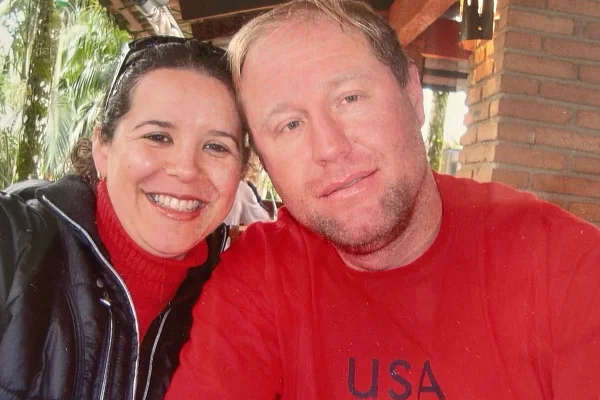 Foto colorida de casal vestindo blusas vermelhas, em ambiente aberto, sorrindo para a foto - Metrópoles.