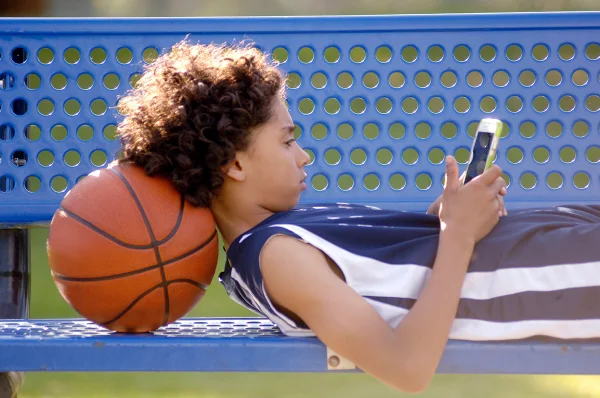 imagem colorida mostra menino usando celular. ele está deitado, com a cabeça apoiada em uma bola de basquete. o menino é negro, tem o cabelo cacheado e usa uma regata com as cores azul e branco. o celular está na mão direita e é da cor branca. metrópoles