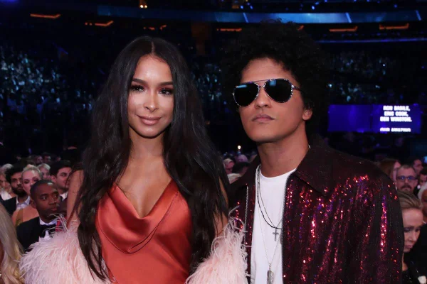 Bruno Mars e Jessica Caban em evento, usando roupas sociais. Cantor aparece de óculos escuros - Metrópoles