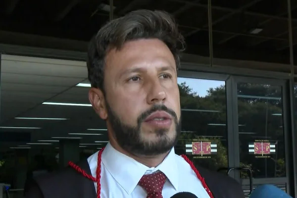 Imagem colorida mostra homem branco com cabelos escuros e barba feita. Ele veste camisa branca e gravata vermelha e uma capa preta de promotor do MInistério Público - Metrópoles
