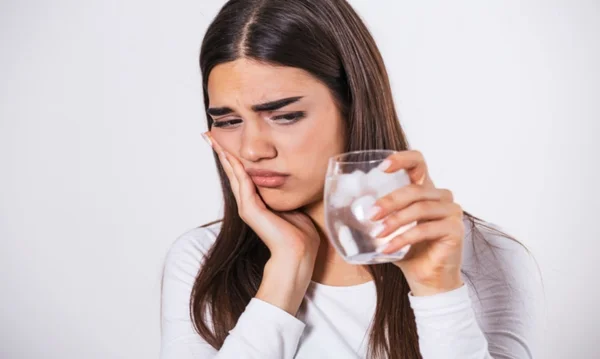 Foto mostra mulher com expressão de desconforto, segurando os dentes enquanto olha um copo de água gelada