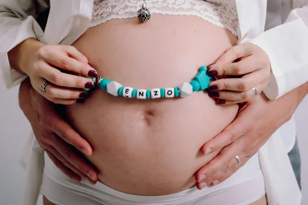 Barriga de grávida com as mãos do pai abraçando e as da mãe segurando miçangas com o nome Enzo