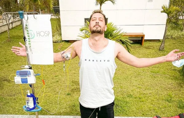 Em foto colorida jovem de camiseta regata branca matém braços abertos, com a cabeça voltada para o céu, de olhos fechados, em frente a um gramado. Ele está com soro no braço - Metrópoles