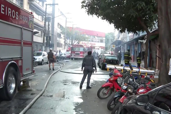Imagem colorida mostra, à esquerda, um caminhão do Corpo de Bombeiros estacionado em uma rua. Ao lado há uma mangueira estendida no chão da rua. No lado direito, há 3 motos estacionadas perto da calçada. Bombeiros caminham pela rua - Metrópoles