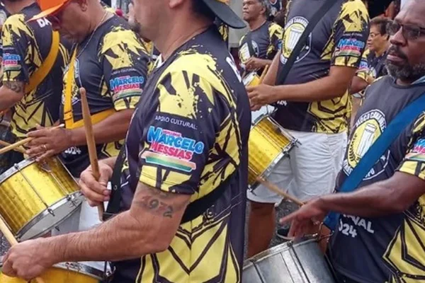 Imagem mostra ritmista do bloco Atrás do Copo com camisa do bloco e propaganda do vereador Marcelo Messias na manga