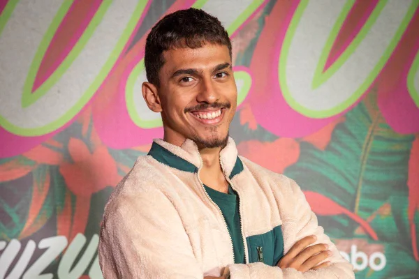 foto de homem moreno de bigode vestindo casaco bege e sorrindo de braços cruzados - metrópoles