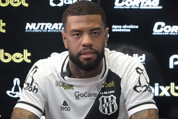 Junior Tavares, lateral-esquerdo da Ponte Preta e ex-São Paulo foi diagnosticado com tumor no cérebro. Ele é um homem negro, jovem, com barba e cabelos pretos, e está vestido com uma camisa da Ponte Preta, branca com uma faixa preta transversal - Metrópoles